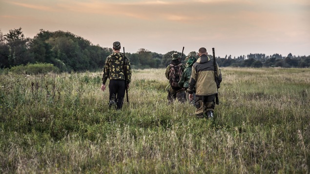 Des chasseurs, de dos, parcourent une prairie, leurs fusils accrochés sur le dos.