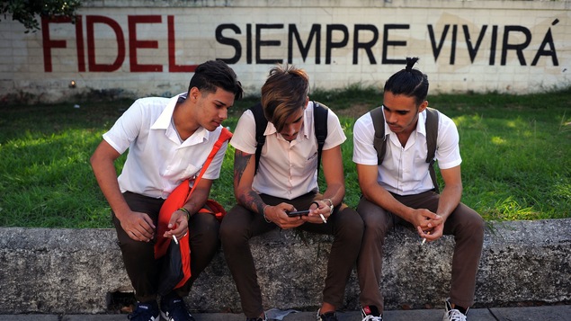 Des jeunes Cubains regardent un téléphone intelligent, assis devant une affiche de propagande à La Havane.