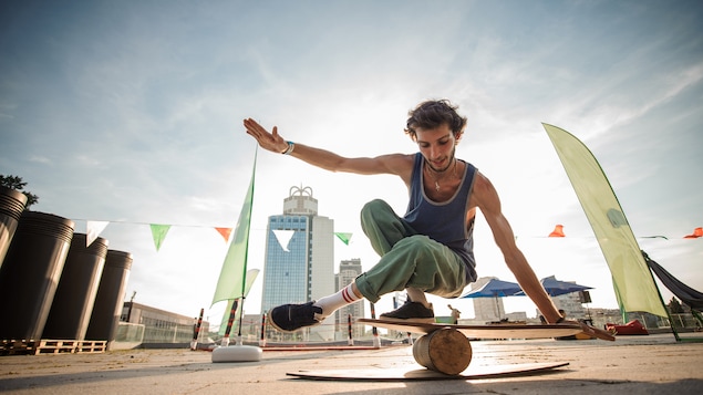 Un jeune acrobate se tient en équilibre sur une bûche de bois surmontée d'une planche.