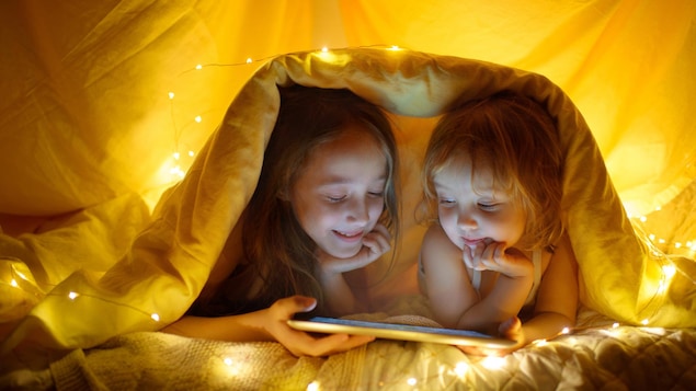 Deux enfants sous une couverture regardent un film sur une tablette.