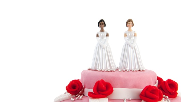 Des figurines représentent un couple de femmes sur un gâteau de mariage rose décoré de fleurs.