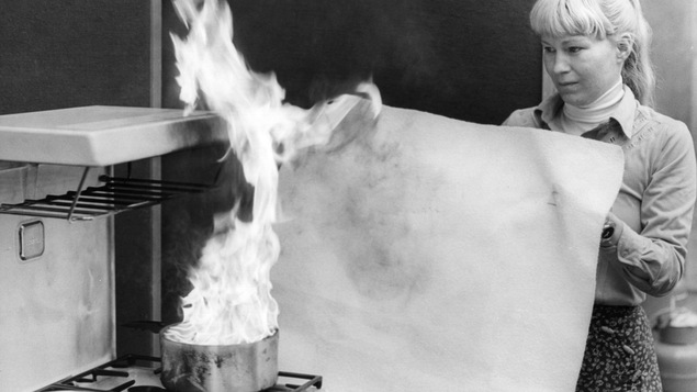 Photo en noir et blanc d'une femme tendant un morceau de tissu à côté d'une flamme sur un poêle.