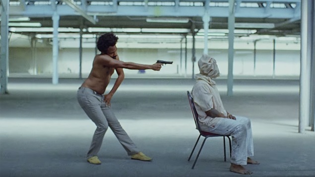 L'artiste Childish Gambino tient un pistolet devant un homme à la tête couverte d'un sac, assis sur une chaise, dans cette omage tirée du vidéoclip de la chanson <i>This Is America</i>.