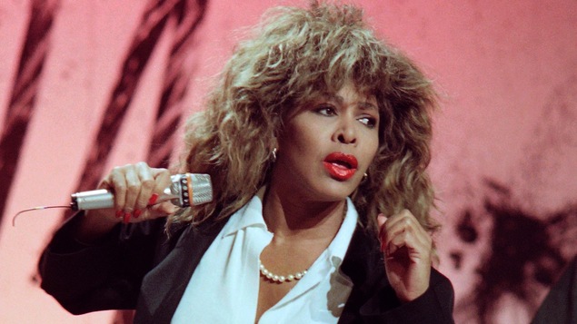 Tina Turner en mouvement sur scène, un microphone à la main.