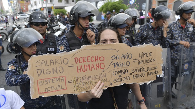 Sous le regard de la police, une travailleuse de l'État tient une affiche réclamant de meilleurs salaires lors d'une manifestation au Venezuela.