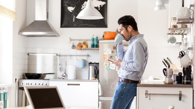 Dans une cuisine, un homme qui a des écouteurs dans les oreilles boit dans une tasse.