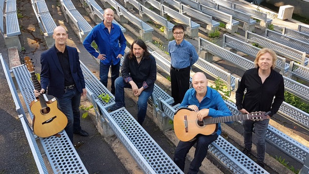 Six hommes sont photographiés plan en plongé dans les estrades, à l'extérieur, et deux des six hommes tiennent une guitare à la main.