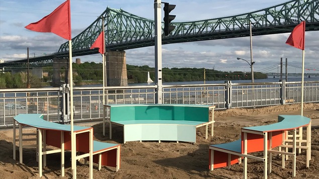 Photo du village au Pied-du-Courant, avec des bancs colorés sur un parterre de sable et le pont Jacques-Cartier en arrière-plan.