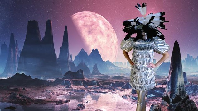 Un mannequin avec un costume rappelant les vêtements traditionnels autochtones, sur fond de paysage lunaire dans des teintes pastel.