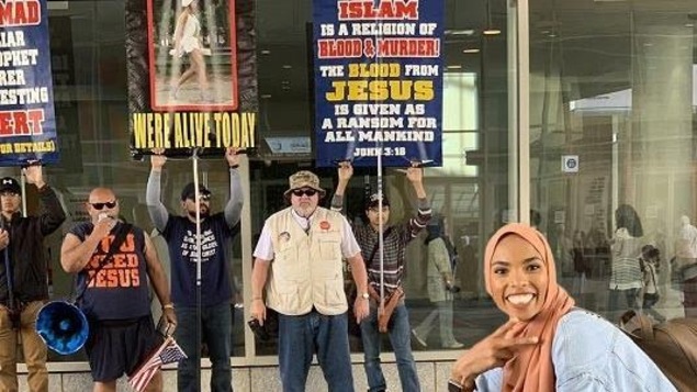 Une jeune femme avec un hijab sourit devant des manifestants chrétiens tenant des pancartes anti-Islam.