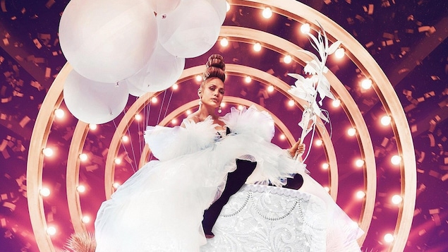 Jennifer Lopez sur un gateau géant avec des ballons blancs, habillée d'une robe de tulle, sur un arrière fond rose et mauve de lumières et de confettis.