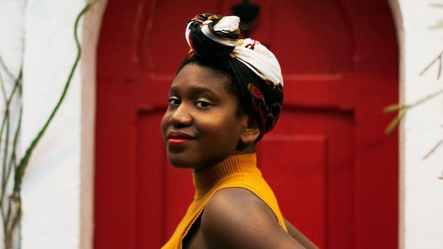 Portrait de Jessica Beauplat avec un foulard sur la tête. Elle regarde la caméra à l'oblique devant une porte rouge.