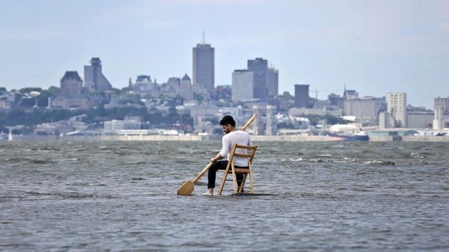 Un homme est assis sur une chaise avec une rame au milieu de l'eau, avec la ville de Québec en arrière-plan.