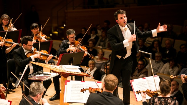Adam Johson dirigeant l'Orchestre symphonique de Montréal et 5 violonistes sur scène.