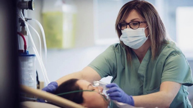Une infirmière qui porte le masque sanitaire ajuste le masque d’un respirateur artificiel sur le visage d’une patiente couché dans un lit d’hôpital. 