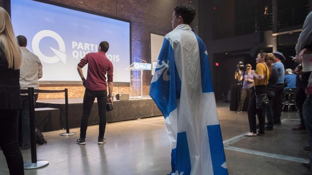 Lors des élections québécoises de 2018, des partisans sont rassemblés au siège du Parti québécois; un des partisans est drapé du drapeau québécois.