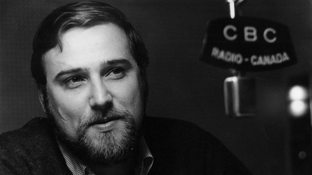 Une photo en noir et blanc d'un homme qui porte une barbe et qui sourit à un micro de Radio-Canada.