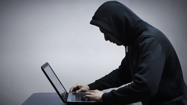 Un homme au visage caché sous le capuchon de son chandail noir s'affaire sur un ordinateur portable.