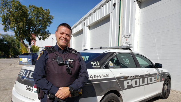Martin Boileau, policier au Service de police de Winnipeg, agent de liaison aux écoles. Habillé en tenue de policier et debout devant un auto de police.