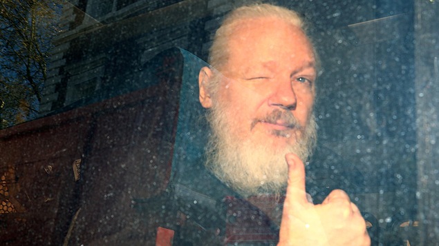 Julian Assange assis à l'arrière d'une voiture, faisant un clin d'oeil à la caméra, le pouce levé.