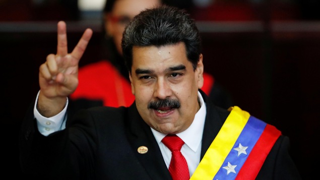 Nicolas Maduro en costume cravate fait un signe de paix avec sa main droite. Il arbore un dossard aux couleurs du drapeau vénézuélien: le jaune, le bleu et le rouge.