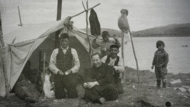 Le père Alexis Joveneau avec une famille innue. Ils sont tous assis près d'une tente, sur le bord de l'eau.
