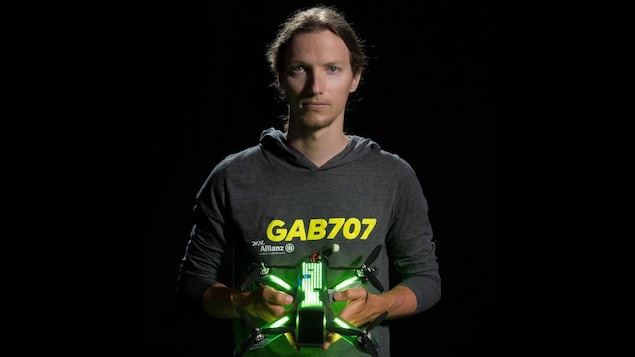 Une photo de Gabriel Kocher habillé d'un chandail gris à capuchon sur lequel est inscrit GAB707 en jaune. Il tient un drone à quatre hélices dans ses mains. Le dessus du drone est éclairé grâce à des dizaines de lumières LED vertes.