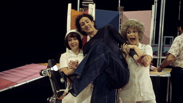 Dans un studio de télévision, les comédiennes Francine Ruel et Dorothée Berryman sont déguisées en infirmières et soutiennent le comédien Robert Gravel qui chausse des patins à roulettes.