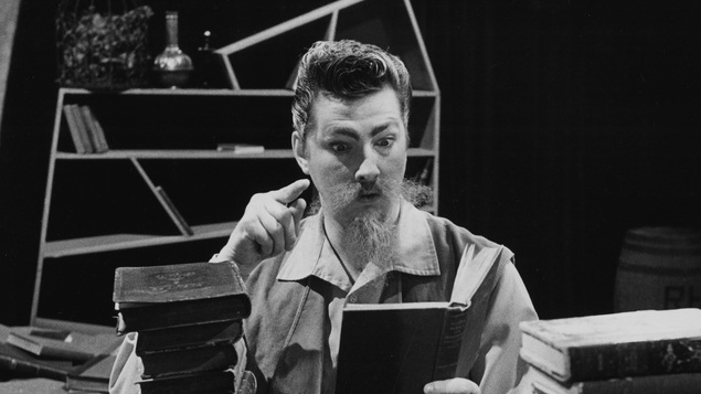 Assis à une table de travail, Robert Gravel dans le rôle de Don Quichotte consulte un de ses bouquins empilés sur son bureau