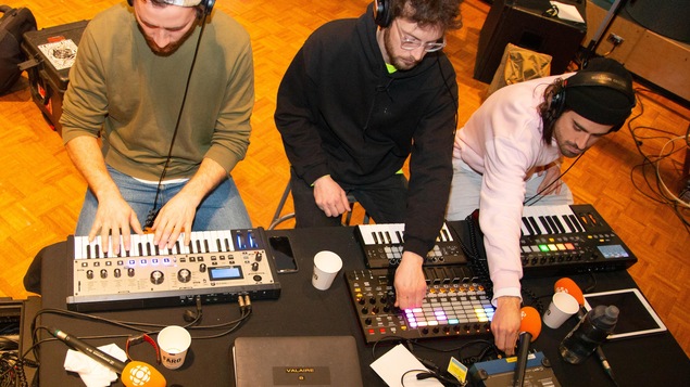 Trois hommes sont assis derrière une table sur laquelle sont étalés des claviers et autres appareils électroniques pour musiciens.