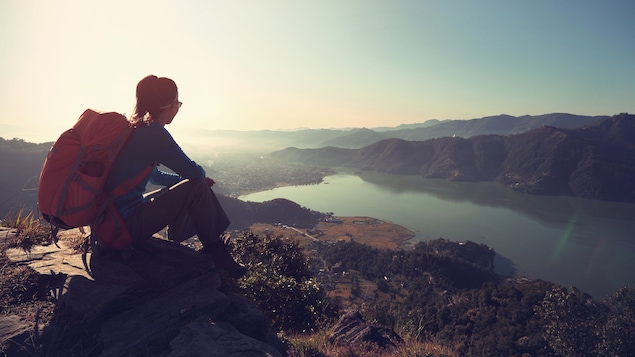 Assise sur un sommet, une femme avec un sac à dos regarde une étendue d'eau.