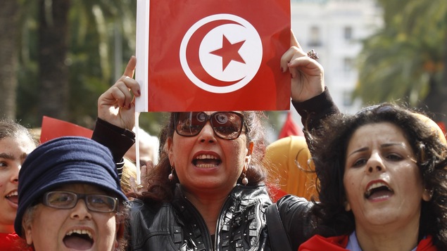 Une femme tient dans ses mains le drapeau tunisien. Le drapeau est rouge et blanc. À ses côtés, deux femmes portent du rouge.