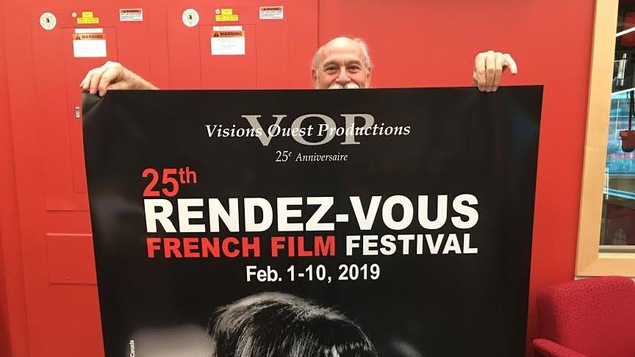 Un homme se tient derrière une affiche qu'il tient et qui annonce les 25e Rendez-vous du cinéma québécois et francophone.