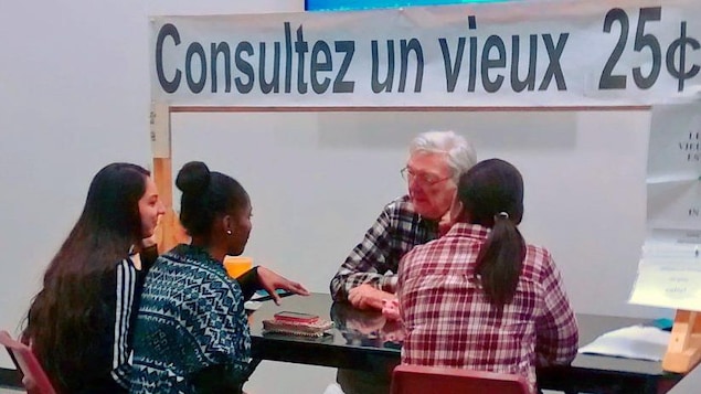Un homme âgé assis à un kiosque devant trois étudiantes.