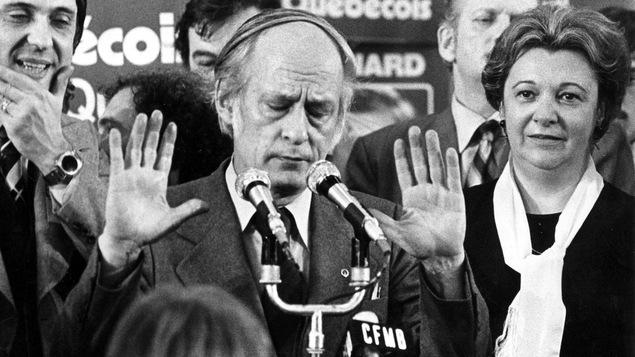 René Lévesque, entouré de candidats élus, ferme les yeux en levant les deux bras vers la foule pour tenter de la calmer.