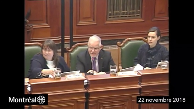 Un homme parle dans un micro. Il est assis entre deux femmes qui ont visiblement l'air choquées.