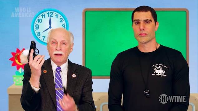 Philip Van Cleave tient un pistolet à la main orné d'une peluche pour enfant dans un décor de classe pré-scolaire. À droite, Sacha Baron Cohen est déguisé en israélien musclé, un sifflet dans le cou.
