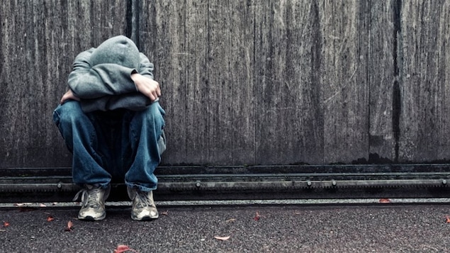 Un jeune adulte recroquevillé sur lui-même, seul devant un grand mur grisâtre.