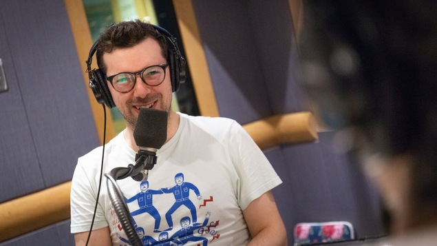 Une photo de Simon Boulerice, qui porte des lunettes rondes et un t-shirt blanc sur lequel sont imprimés des personnages bleus.