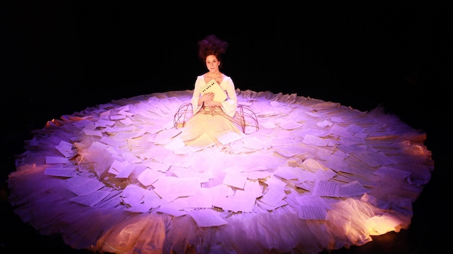 La comédienne Sofi Lambert, assise à genoux sur scène, tient un petit instrument de musique contre sa poitrine. Elle porte une immense robe sur laquelle il y a plusieurs feuilles de papier.