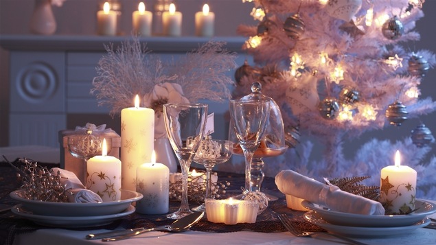 Une table dressée pour Noël dans des couleurs blanches et argentées avec des bougies et un sapin qui brille en arrière-plan.