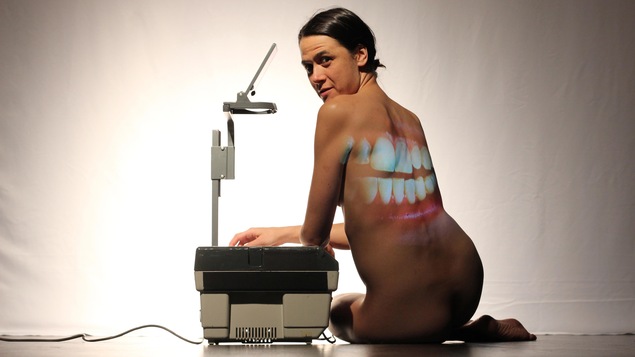 Une bouche est projetée sur le dos d'une femme nue, qui est assise à côté d'un rétroprojecteur.