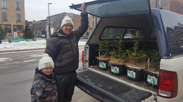 Un père et son fils sourient à la caméra devant une voiture remplie de sapins baumiers.