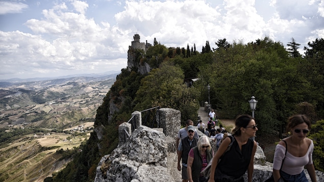 Des touristes marchent sur une route escarpée dans la montagne. En arrière-plan, la tour d'un château.