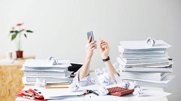Photo conceptuelle où des bras sont visibles derrière un bureau rempli de piles de papiers.