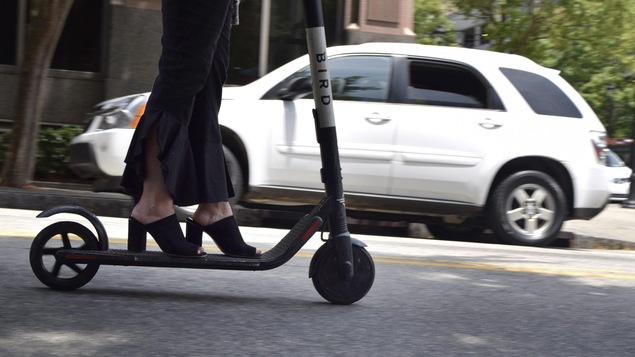 Une femme portant des chaussures à talons conduit une trottinette électrique sur une rue.