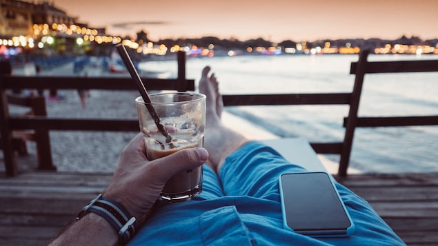 Un touriste savoure un breuvage à la plage avec son téléphone cellulaire posé sur la cuisse.