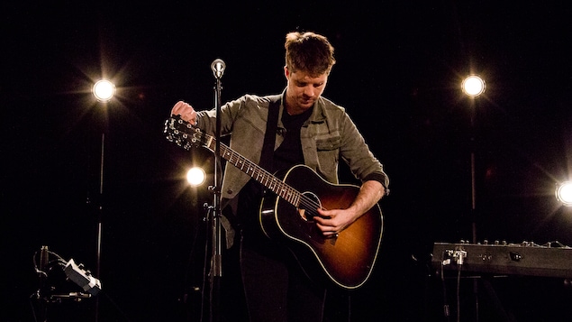 Vincent Vallières accorde sa guitare au Studio 12. Micro devant lui, quatre projecteurs se trouvent derrière lui. Le fond de la photo est noir.