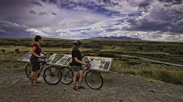 Deux cyclistes debout avec leurs vélos devant des plaques explicatives, avec une rivière, une plaine et des montagnes en arrière-plan.