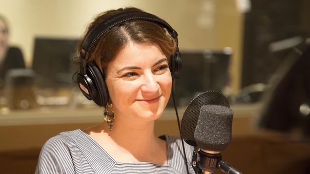 Elle sourit, des écouteurs sur la tête et assise derrière un micro.